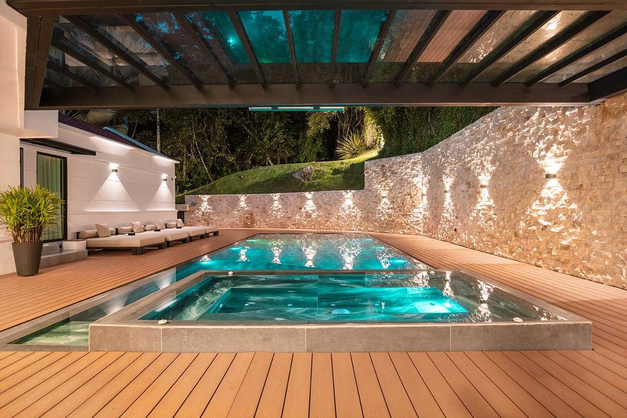 Piscina bajo pérgola con muro en piedra iluminado y deck en madera